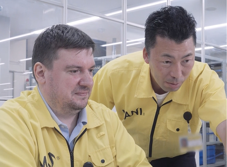 Two men in yellow MANI uniforms working together at a computer in a modern industrial or lab setting.