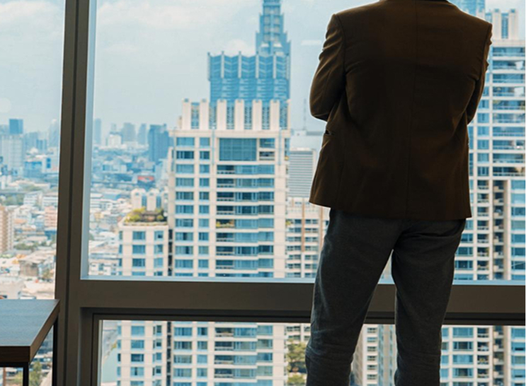 A person in business attire standing by a large window, overlooking a cityscape with tall modern buildings.