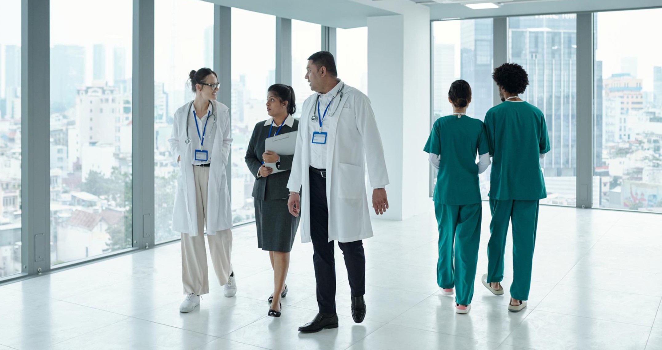 Group of medical professionals walking and talking in a modern hospital hallway with large windows.