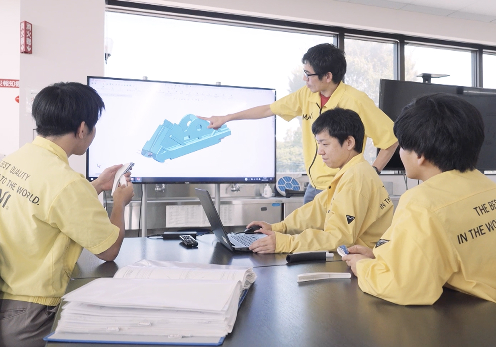 Four engineers in yellow MANI uniforms reviewing a 3D CAD model on a large monitor during a design meeting