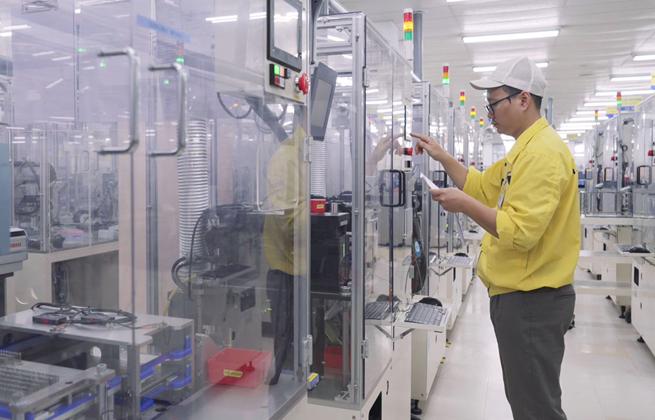A technician in a yellow shirt operates automated machinery inside a clean, high-tech manufacturing facility