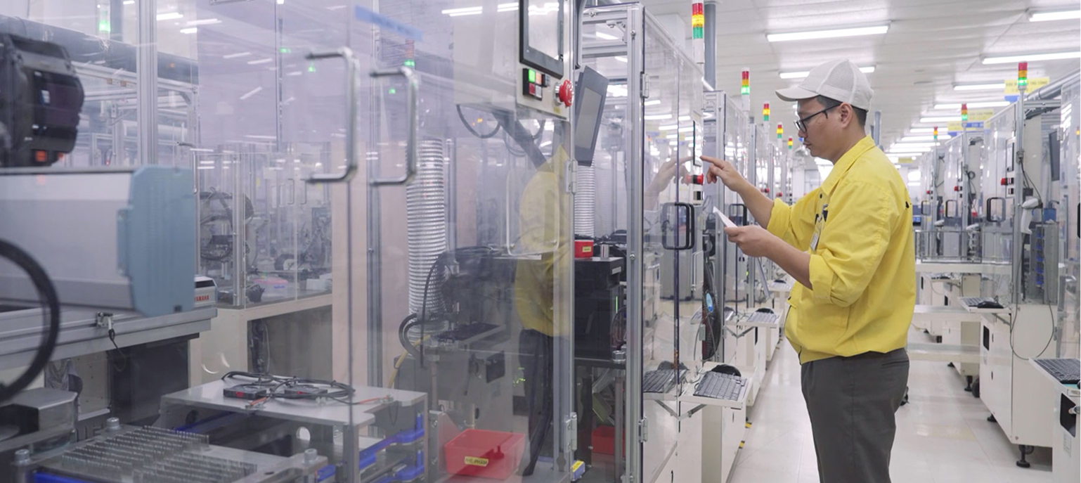 A technician in a yellow shirt operates automated machinery inside a clean, high-tech manufacturing facility