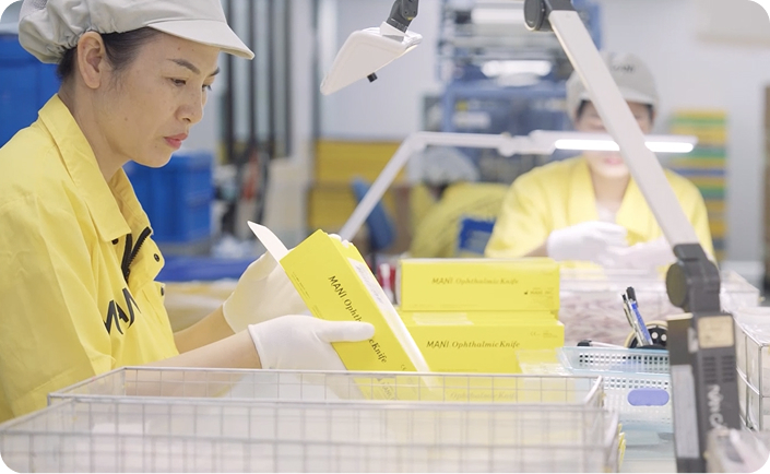 Workers in yellow uniforms inspecting and packaging yellow boxes labeled 'MANI Ophthalmic Knife' at a production station.