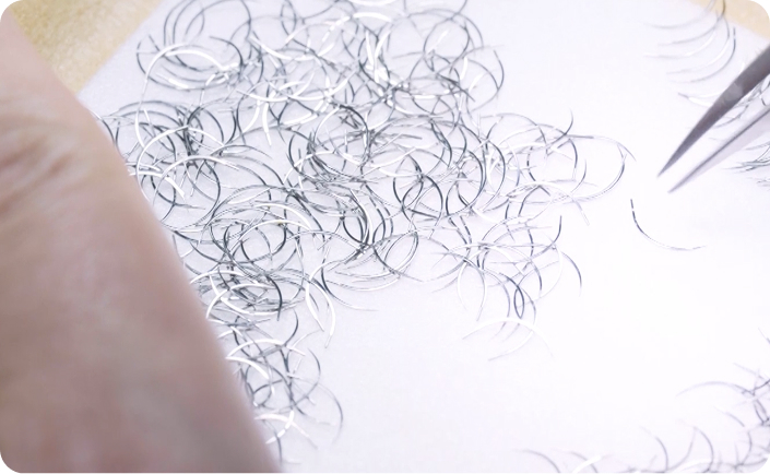 Close-up of curved surgical needles being sorted or inspected with tweezers on a white surface.