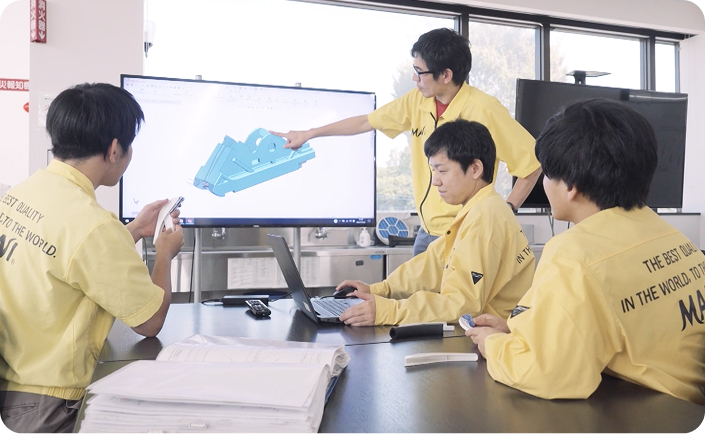 Four engineers in yellow shirts are discussing a 3D CAD model displayed on a large screen in a bright office environment.
