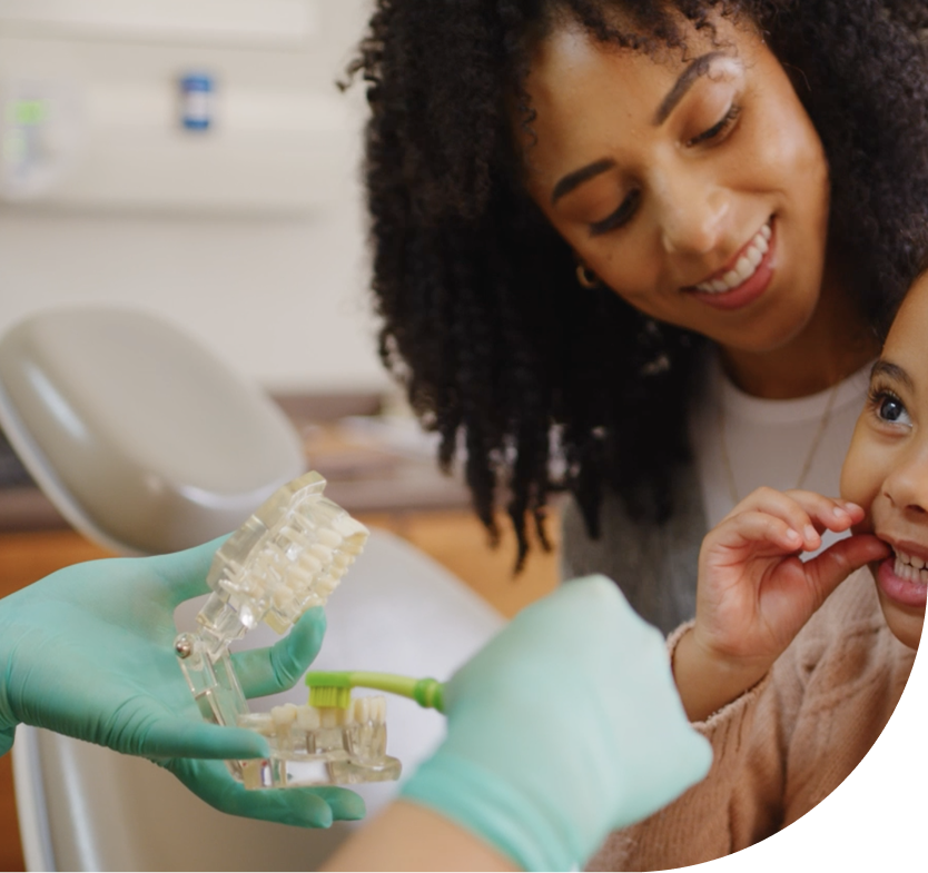 A dentist or hygienist teaching a child how to brush teeth using a dental model, while a smiling woman looks on supportively.